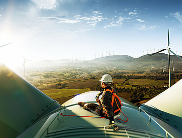 Ein glücklicher Ingenieur, der sich nach gelungener Arbeit über seinen Erfolg freut. Er steht oben auf einer Windkraftanlage und blickt auf eine schöne Sonnenuntergangslandschaft.