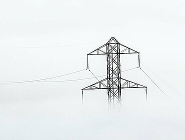 Stromleitungen ziehen sich durch dichten Nebel. Metallene Strommasten durchstoßen die neblige Landschaft und tragen unsichtbar die Elektrizität.