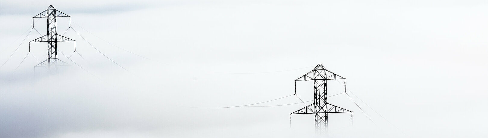 Fog-shrouded power lines. Stromleitungen ziehen sich durch dichten Nebel. Metallene Strommasten durchstoßen die neblige Landschaft und tragen unsichtbar die Elektrizität.